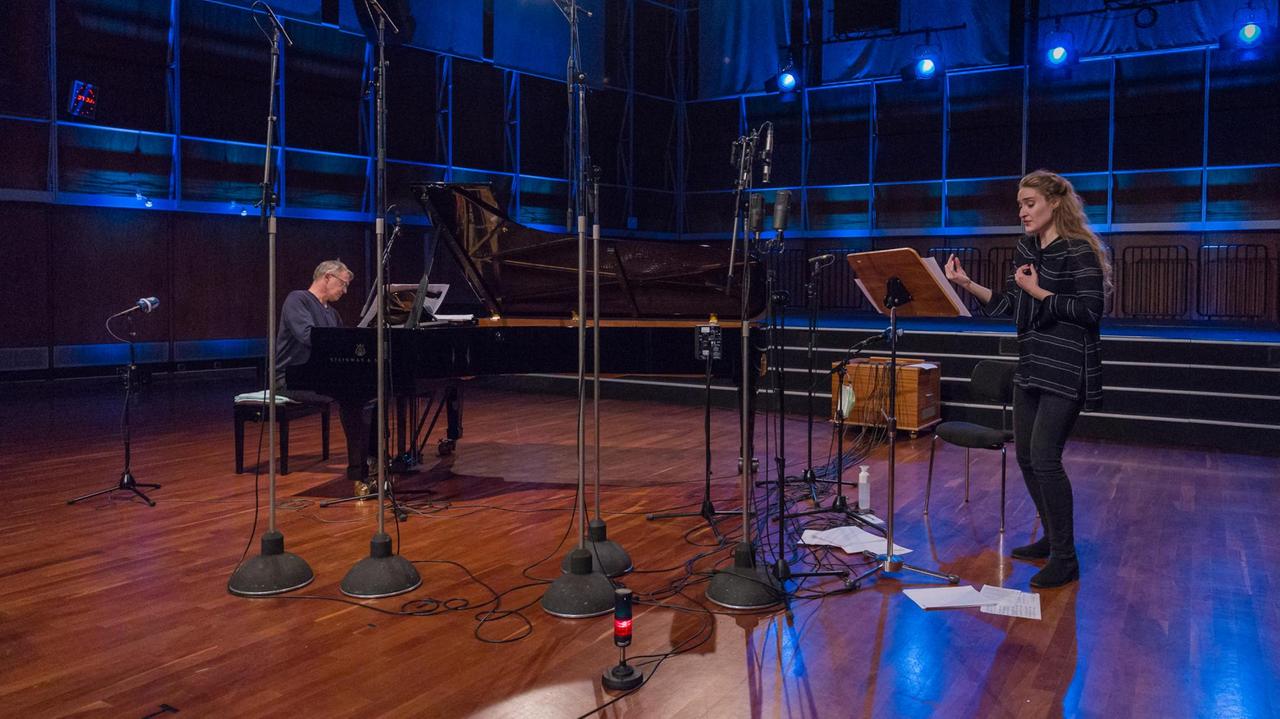 In einem holzgetäfelten Konzertsaal stehen viele Mikrofone, rechts im Bild ist eine junge Sängerin zu sehen, links ein Pianist am schwarzen Flügel.