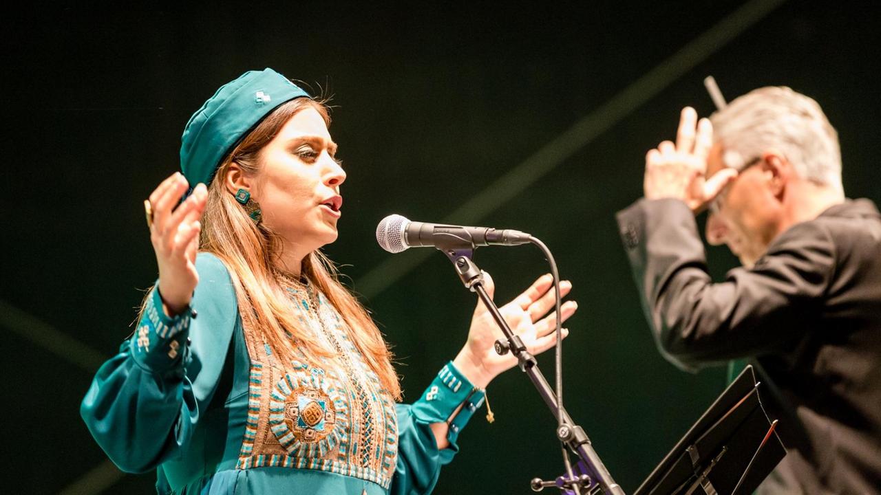 Eine Frau steht auf einer Bühne und singt in ein Mikrofon, rechts neben ihr steht der Dirigent und hält seine linke Hand in Kopfhöhe.