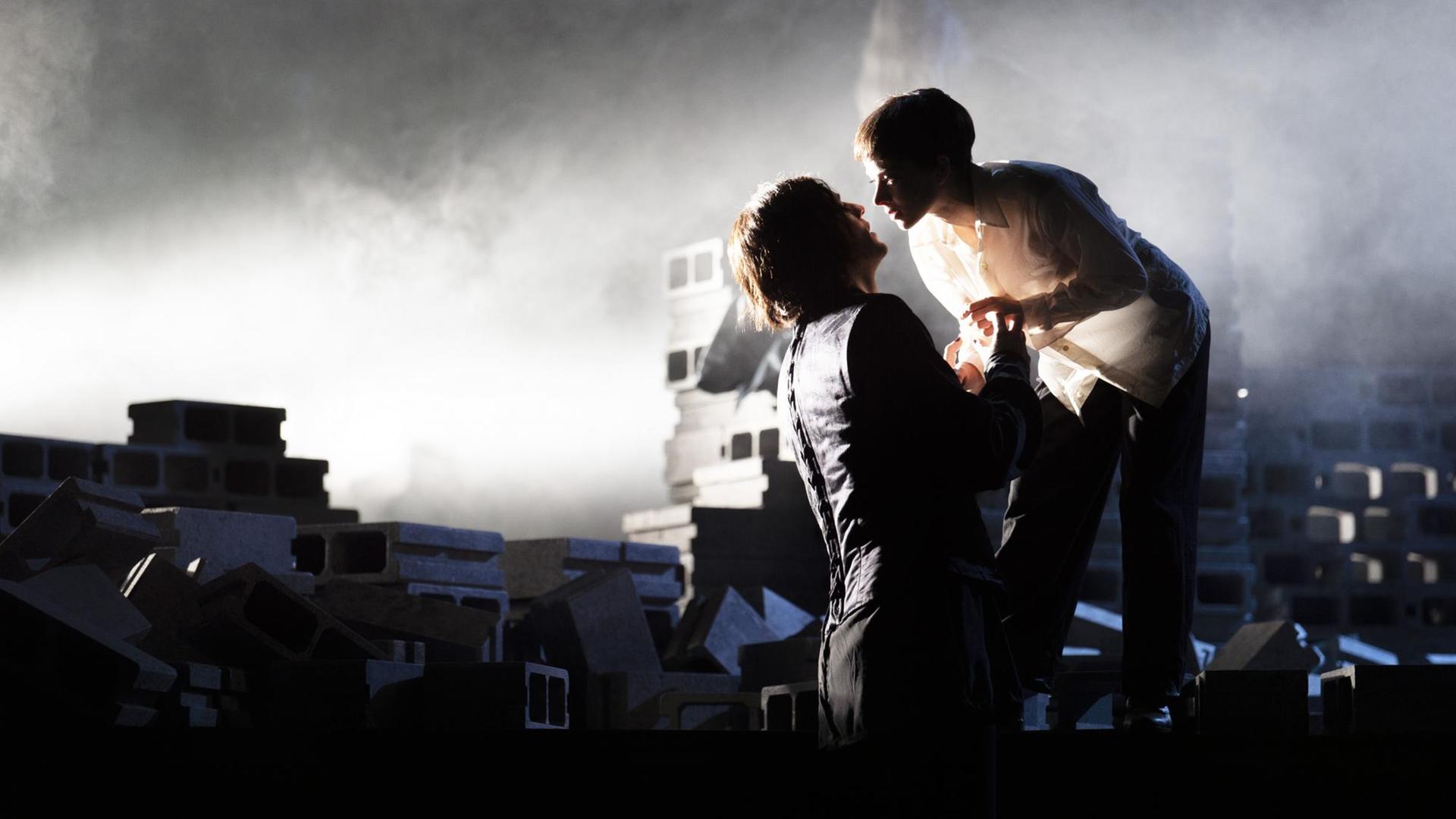 Auf einer Bühne, auf der sich Steine stapeln, stehen zwei Personen, ihre Gesichter zueinander geneigt, als würden sie sich gleich küssen.