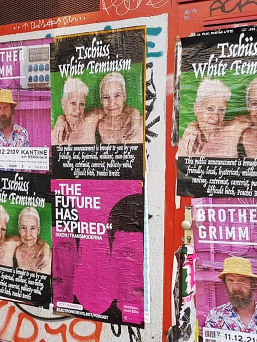 An der Wand sind viele gleiche Plakate zu sehen. Das Motiv zeigt zwei unbekleidete Frauen, zu sehen bis kurz vor Brusthöhe, die sich umarmen. Über ihren Köpfen steht "Tschüss, White Feminism"