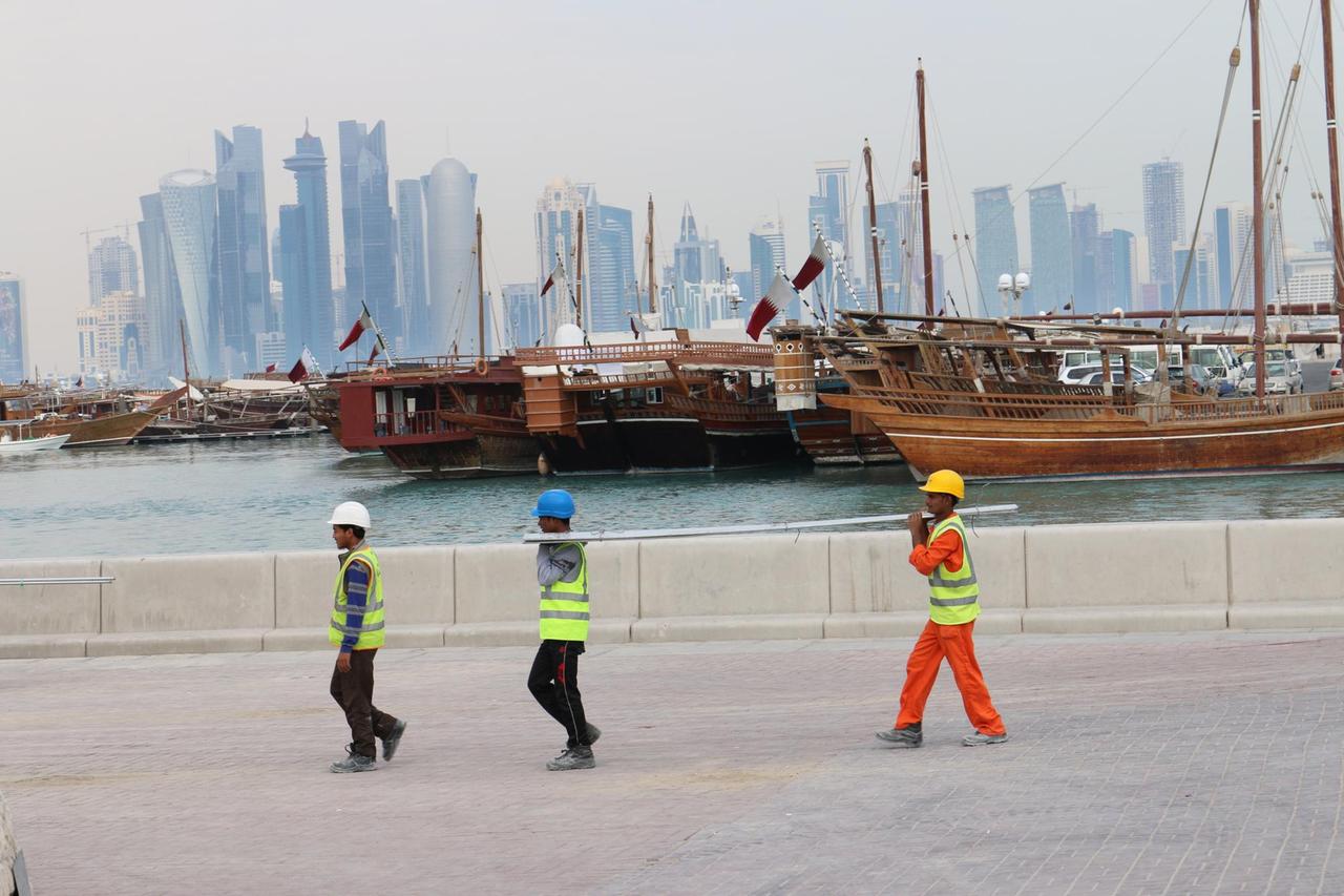 Drei Bauarbeiter gehen am Dau-Hafen entlang mit der Skyline der katarischen Hauptstadt Doha im Hintergrund.