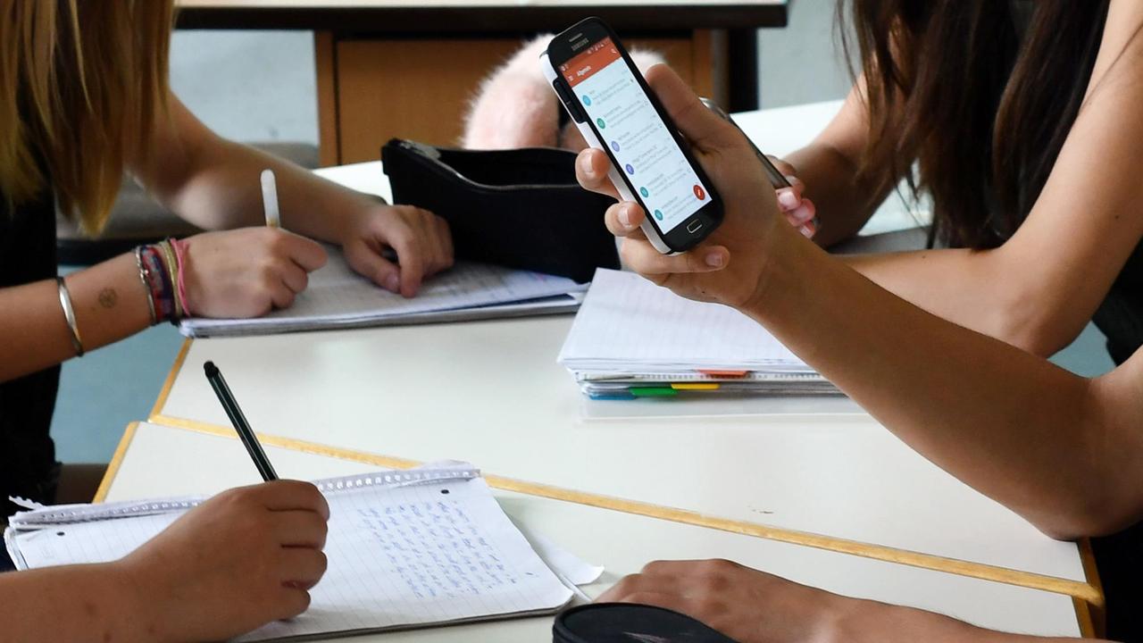 Die Hand eines Schülers oder einer Schülerin hält ein Handy hoch. Dahinter die Arme von weiteren Schülern, die etwas schreiben. 