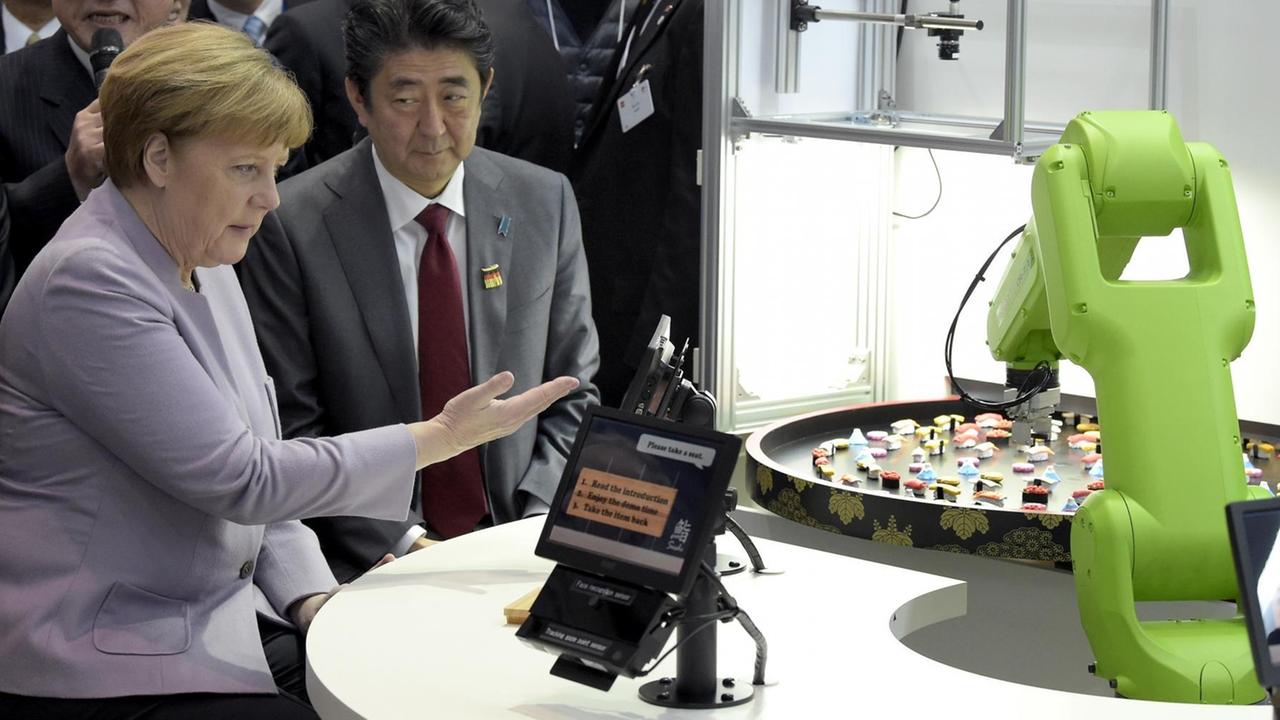 Bundeskanzlerin Angela Merkel und Japans Premier Shinzo Abe sitzen an einem Tisch, vor ihnen ein Tabletcomputer zur Steuerung eines grünen Roboterarms.