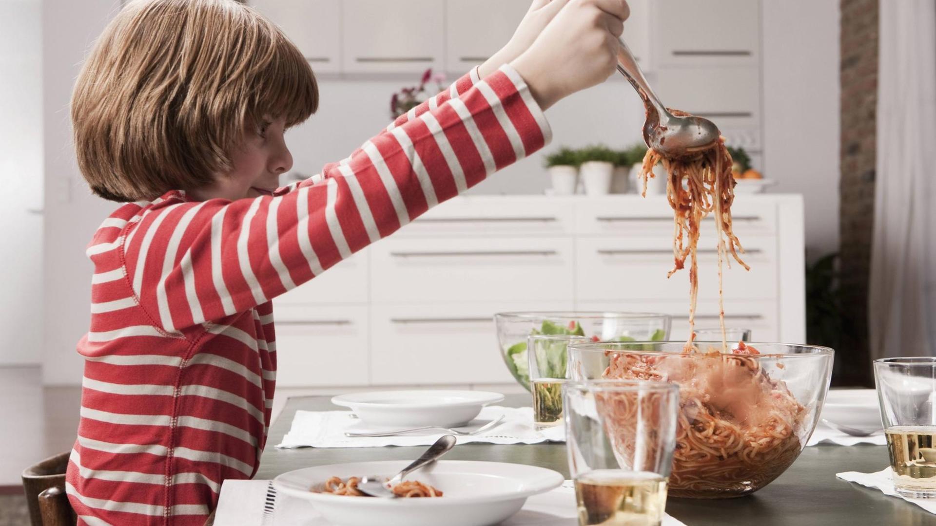 Ein Junge füllt sich Spaghetti mit Tomatensoße aus einer Schüssel auf seinen Teller