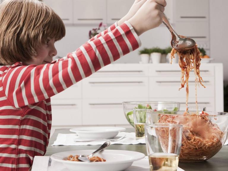 Ein Junge füllt sich Spaghetti mit Tomatensoße aus einer Schüssel auf seinen Teller