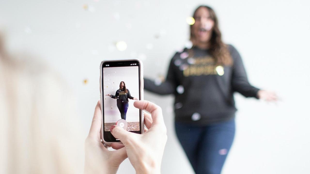 Eine Frau posiert mit Konfetti vor einer Kamera, sie ist doppelt zu sehen, auf dem Bildschirm und im Hintergrund des Bildes.