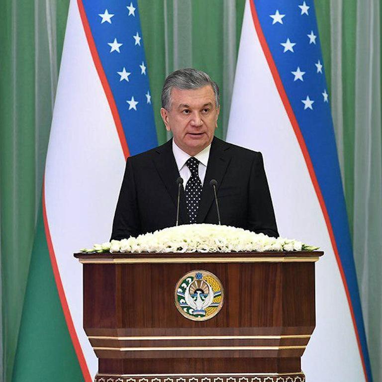 Shavkat Mirziyoyev steht an einem Rednerpult, im Hintergrund mehrere Landesflaggen.