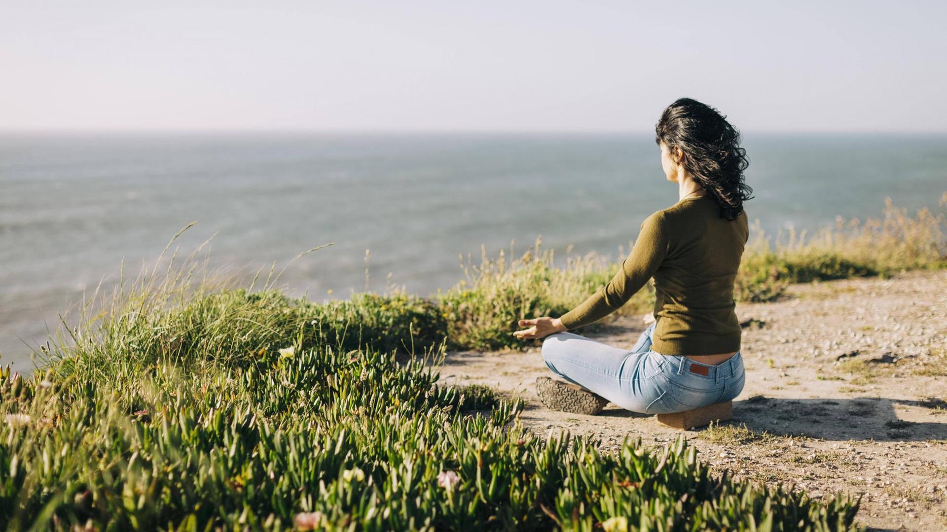 Eine Frau sitzt meditierend im Lotussitz auf dem Boden. Im Hintergrund sieht man das Meer.