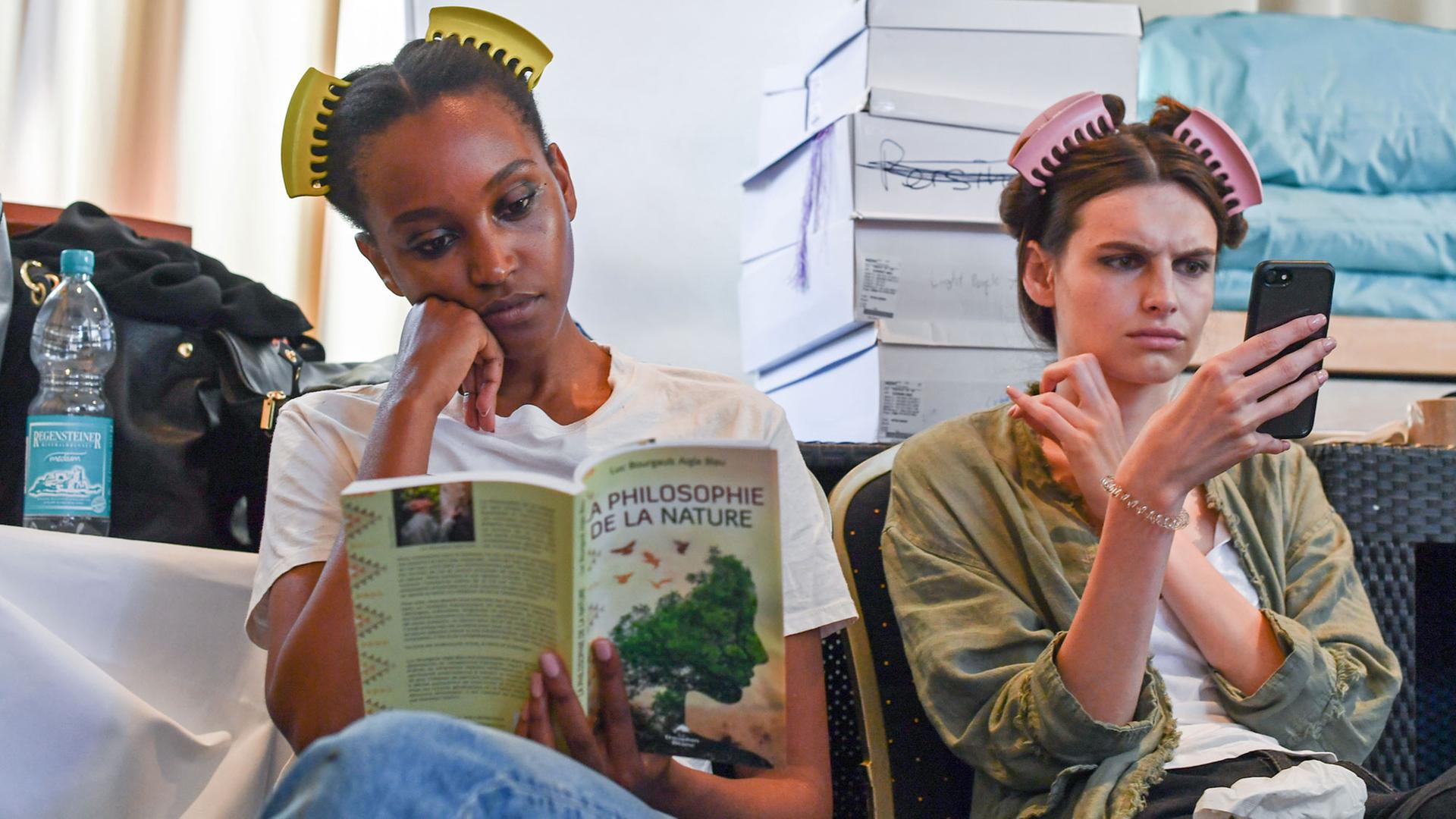 04.07.2018, Berlin: Models lesen vor der Show des Labels Marcel Ostertag backstage auf der Fashion Week in einem Buch und auf dem Smartphone.