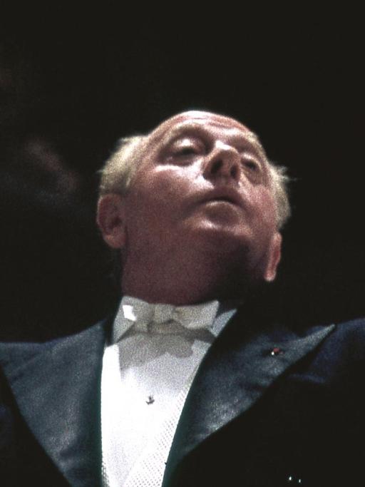 Brustbild des Dirigenten Eugene Ormandy, er dirigiert, der linke Arm ist erhoben, der Blick geht nach oben, er trägt einen schwarzen Konzertfrack mit einem weißen Hemd und einer weißen Fliege, er hat weiße kurze Haare und eine Halbglatze