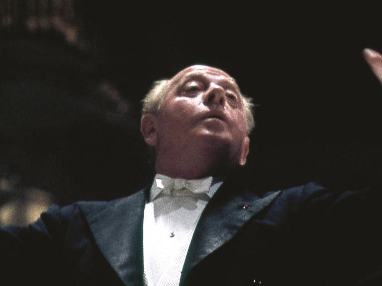 Brustbild des Dirigenten Eugene Ormandy, er dirigiert, der linke Arm ist erhoben, der Blick geht nach oben, er trägt einen schwarzen Konzertfrack mit einem weißen Hemd und einer weißen Fliege, er hat weiße kurze Haare und eine Halbglatze