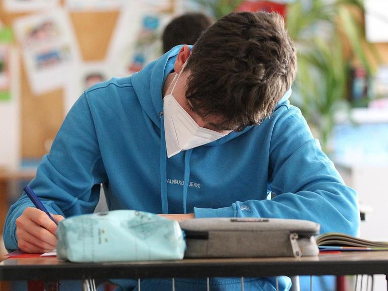 Ein Schüler sitzt mit einem Mundschutz im Klassenzimmer und schreibt. 