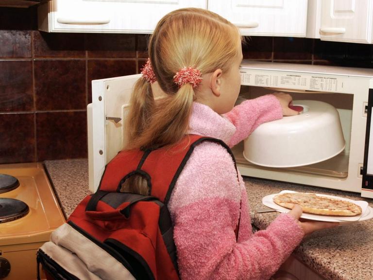 Ein junges Mädchen, das gerade von der Schule nach Haus gekommen ist, wärmt sich in der Mikrowelle etwas zu Essen auf.