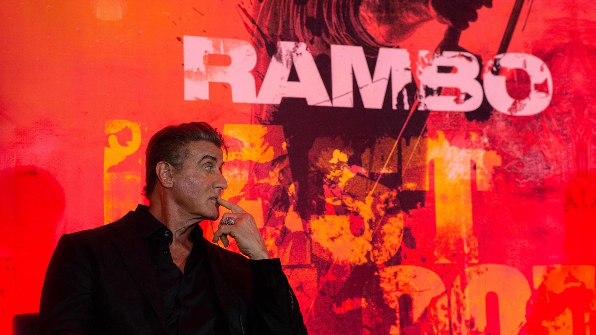 Der Schauspieler Sylvester Stallone sitzt in einem schwarzen Anzug vor einer Wand auf der "Rambo" geschrieben steht.