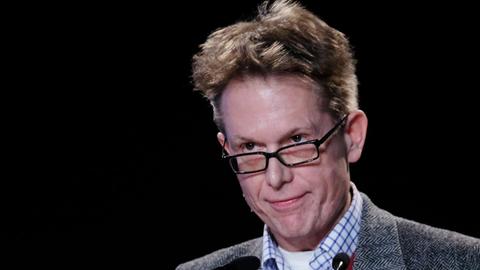 Beck steht mit ernstem Blick hinter zwei Mikrofonen. Er trägt eine Brille. Der Hintergrund ist schwarz.