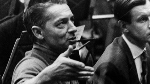 Ein Mann mit Haartolle schaut auf einer Schwarzweißfotografie leicht nach oben und scheint zu reden, dabei hält er seine rechte Hand auf Gesichthöhe.