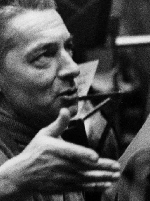 Ein Mann mit Haartolle schaut auf einer Schwarzweißfotografie leicht nach oben und scheint zu reden, dabei hält er seine rechte Hand auf Gesichthöhe.