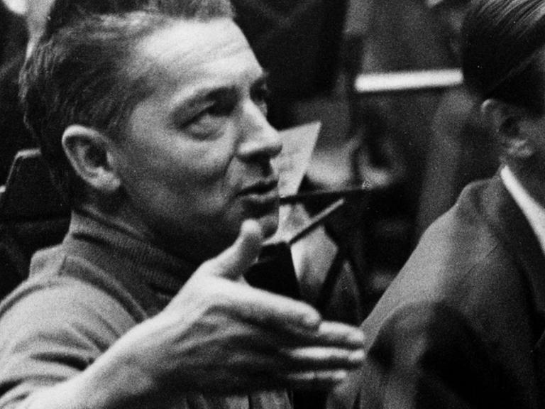 Ein Mann mit Haartolle schaut auf einer Schwarzweißfotografie leicht nach oben und scheint zu reden, dabei hält er seine rechte Hand auf Gesichthöhe.