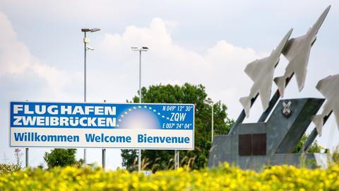Schild mit der Aufschrift "Flughafen Zweibrücken. Willkommen, Welcome, Bienvenue" hinter einer Wiese mit gelben Blumen, daneben eine Skulptur aus drei metallenen Flugzeugen.