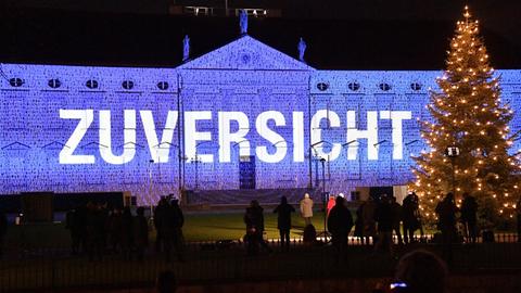 Im Rahmen der Lichtinstallation "Lichtblicke" ist auf das Schloss Bellevue in Berlin in Blau der Schriftzug "Zuversicht" projiziert. Davor steht bei einem Pressetermin unter anderen Bundespräsident Frank-Walter Steinmeier.
