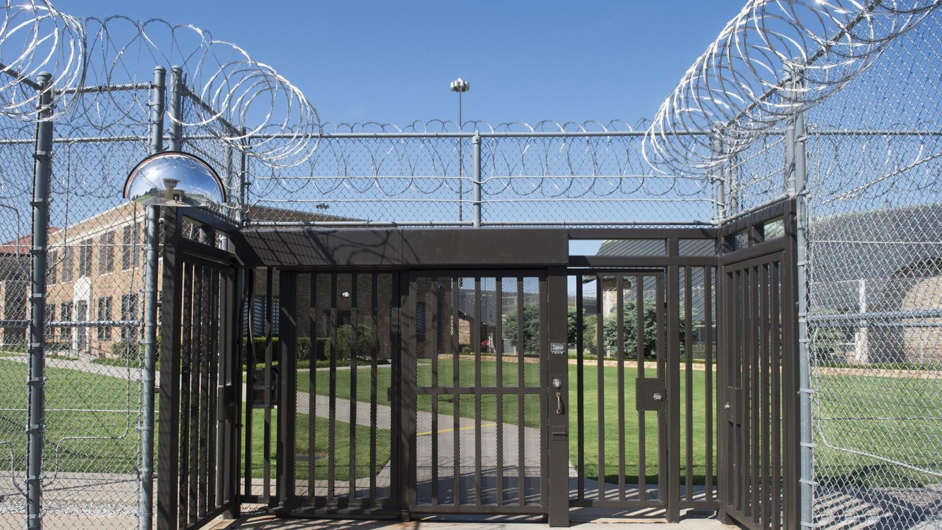 Blick auf den vergitterten Eingang eines Gefängnisses, die Zäune sind mit gerolltem Stacheldraht gesichert