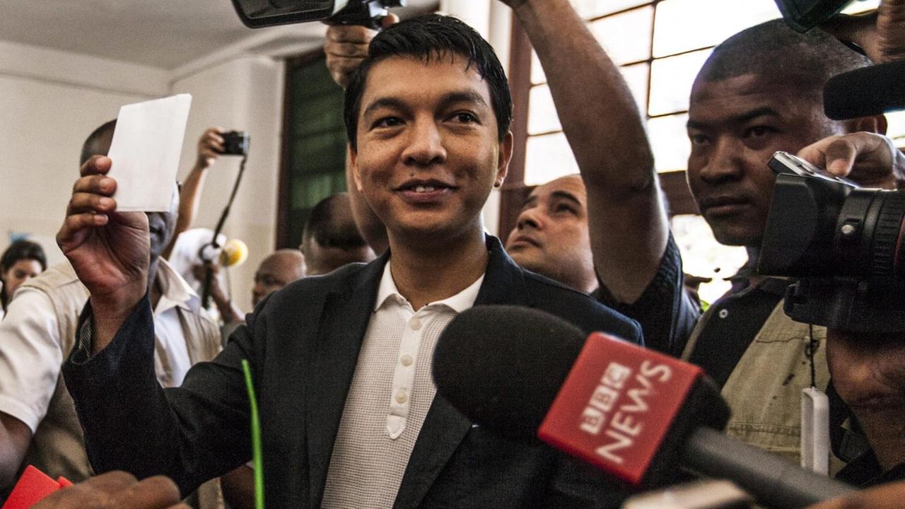 Rajoelina im schwarzen Sakko hält den Stimmzettel lächelnd über die rote Urne. Er ist umgeben von Kameraleuten und Mikrofonen.