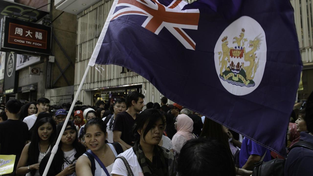 Die Demonstranten marschieren durch eine Straße, jemand hält eine Fahne der ehemaligen Kronkolonie Hongkong.