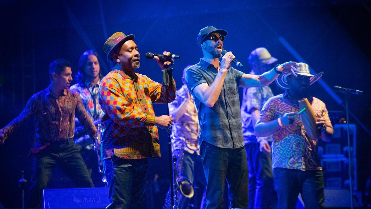 Zwei Männer stehen auf einer Bühne und singen in Mikrofone. Ein Mann trägt ein kariertes Hemd, der zweite ein gelbes Hemd. Um die Männer stehen verschiedene weitere Musiker.
