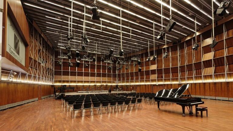 Zu sehen ist der menschenleere Kammermusiksaal im Deutschlandfunk-Gebäude. Ein großer Raum mit Parkett ausgelegt, darauf Stuhlreihen und davor ein Konzertpiano. Von der Decke hängen schwarze Scheinwerfer.