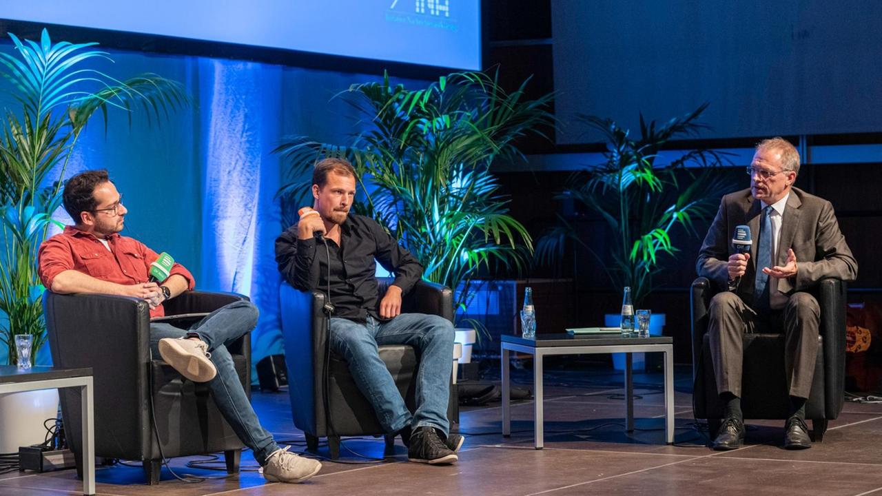 Drei Männer diskutieren auf einer Bühne.