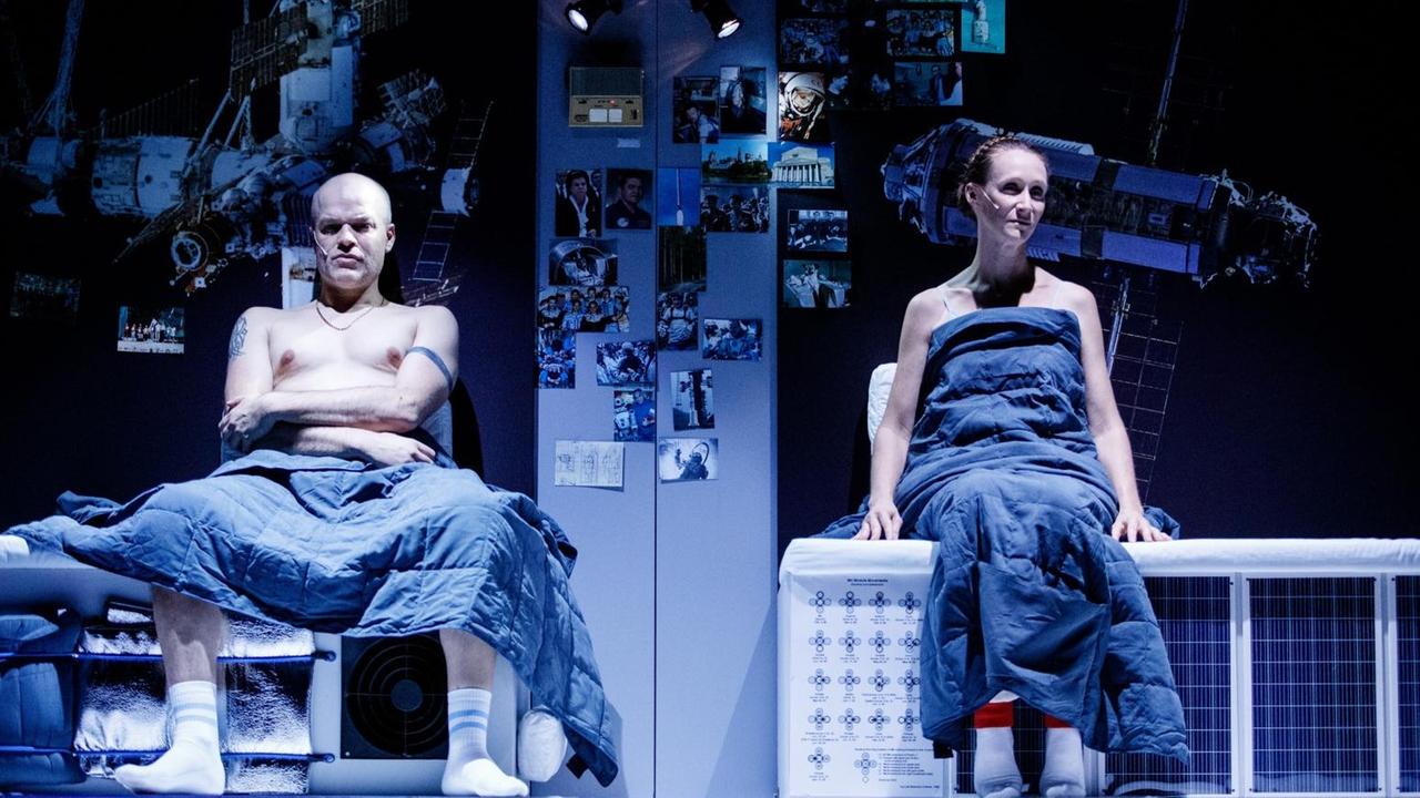 Foto von der Inszenierung: Ein Mann und eine Frau sitzen eingewickelt in Decken auf der Bühne und blicken Richtung Zuschauer.