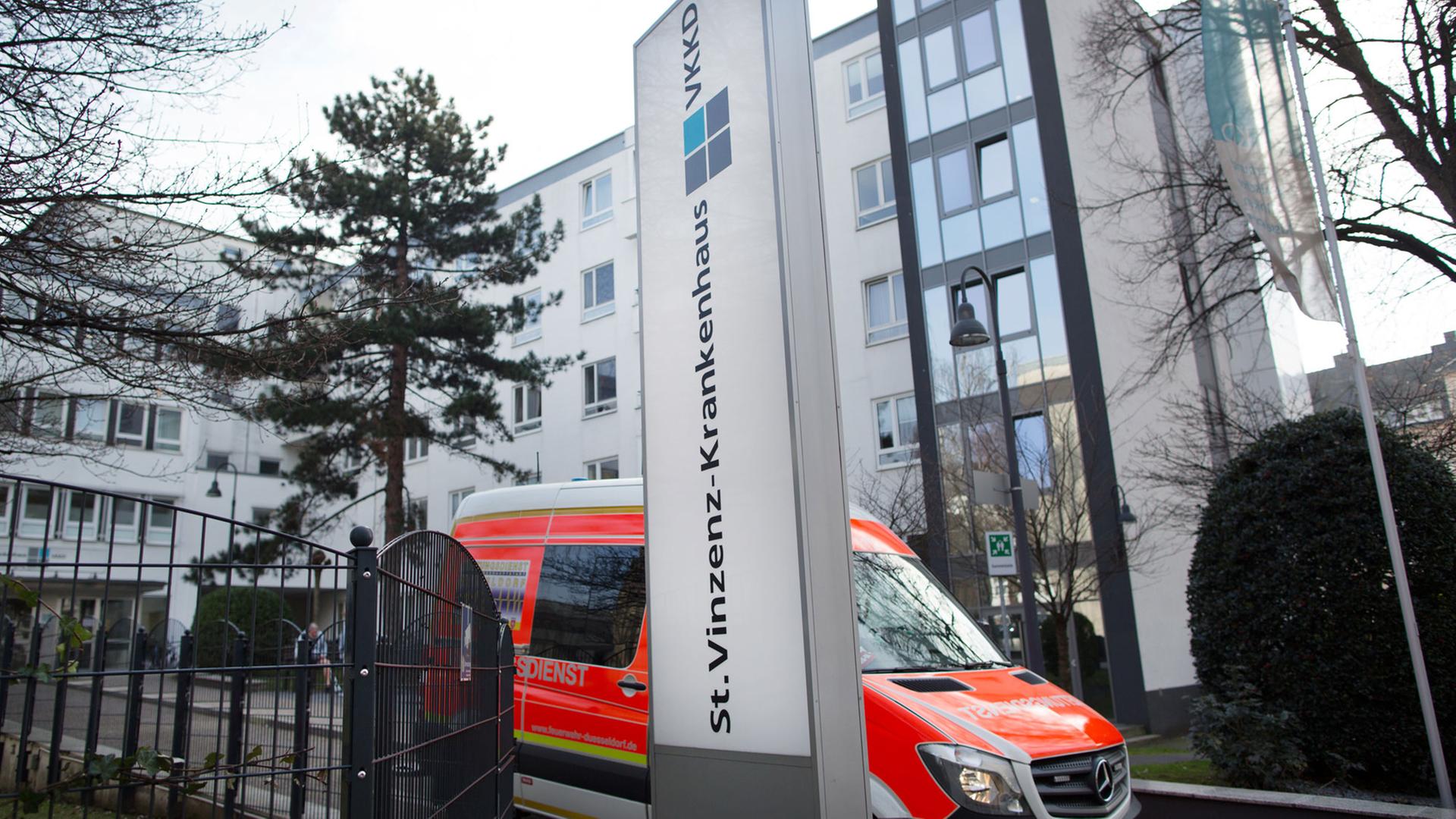 Im Vordergrund ein senkrechtes Schild mit der Aufschrift St.-Vinzenz-Krankenhaus, dahinter fährt ein Rettungswagen vom Klinikgelände, im Hintergrund das Krankenhausgebäude