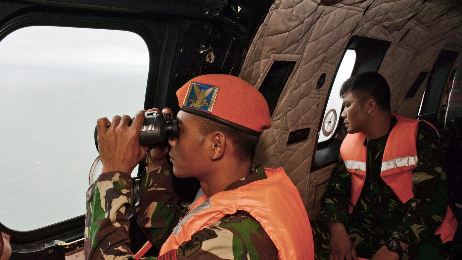 Ein indonesischer Soldat in Uniform und Schwimmweste blickt mit einem Fernrohr aus dem Fenster eines Hubschraubers.