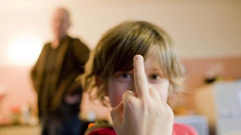 Ein Junge zeigt den Stinkefinger, im Hintergrund ist verschwommen ein erwachsener Mann zu sehen.
