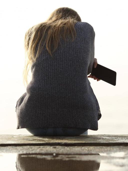 Ein Mädchen sitzt alleine. Ihr Rücken ist dem Betrachter zugewandt. Sie hält ein Smartphone in der Hand.