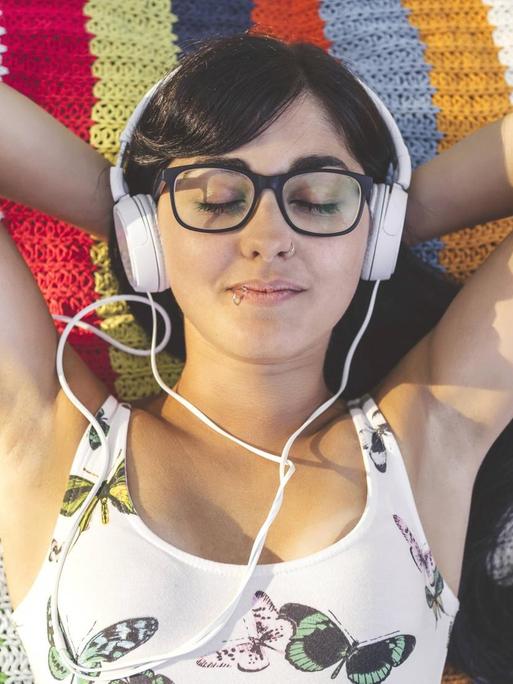 Ein Frau mit Brille liegt auf einer bunten Decke auf einer Wiese, hat Kopfhörer auf und ein Handy neben sich liegen.