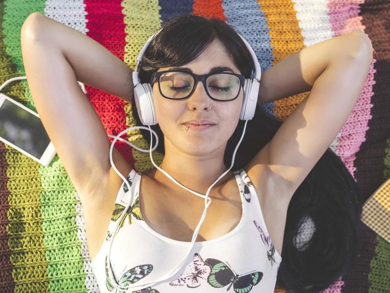 Ein Frau mit Brille liegt auf einer bunten Decke auf einer Wiese, hat Kopfhörer auf und ein Handy neben sich liegen.