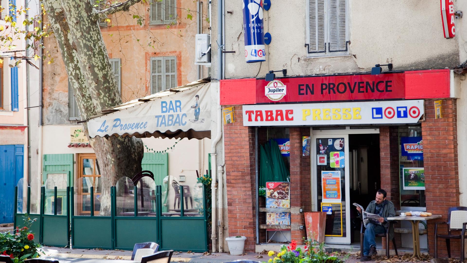 Hauptplatz Place de la Liberation in einem typischen provenzalischen Dorf: Vor der Dorfbar liest ein Mann Zeitung.