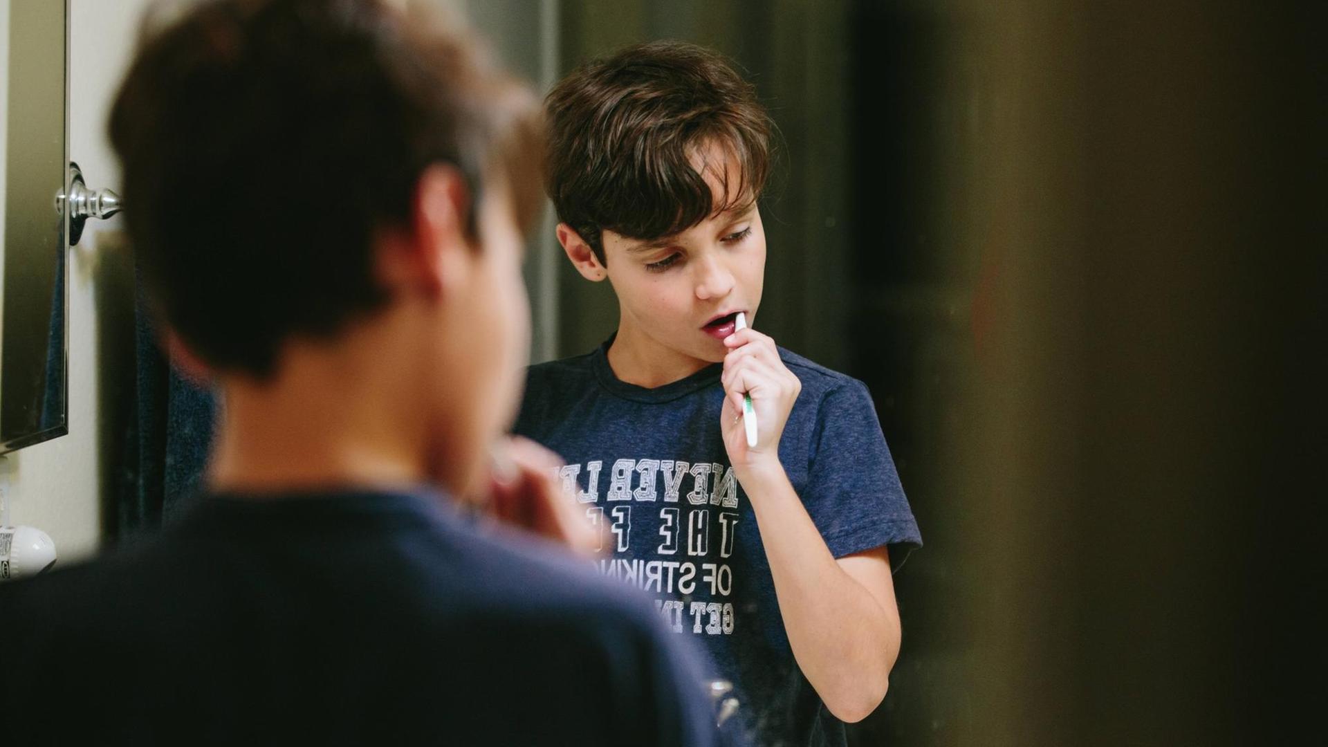 Ein Junge steht vor dem Spiegel und putzt sich die Zähne. Zu sehen sind sein Rücken und im Spiegel seine Reflexion.