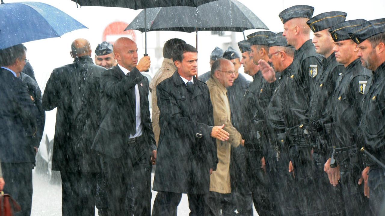 Manuel Valls schüttel die Hand eines Polizisten, der in einer Reihe Polizisten steht. Es regnet, jemand hält Valls einen Schirm über den Kopf.