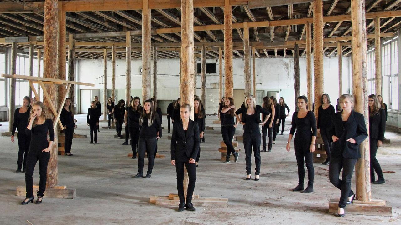 Die Sängerinnen des Chores stehen in einer Halle deren Decke von Holzstützen abgestützt wird.