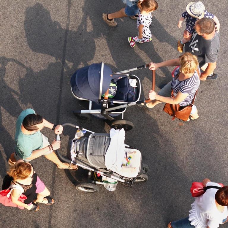 Zwei junge Familien mit Kindern begegenen sich, als sie über eine Straße gehen. Bei der einen Familie schiebt die Mutter den Kinderwagen, bei der anderen der Vater.