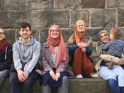 Zwei junge Männer und drei Frauen mit Kopftuch sitzen zusammen auf einer Mauer und schauen in die Kamera. Eine Frau hat ein Kleinkind im Arm.