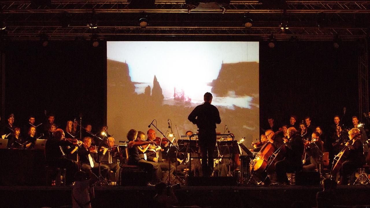 Ein Orchester mit Dirigent sind im Dunklen vor einer Leinwand zu sehen, auf der eine Brandung zu sehen ist. Links und rechts stehen im Hintergrund, neben der Leinwand, Sängerinnen und Sänger eines Chores.