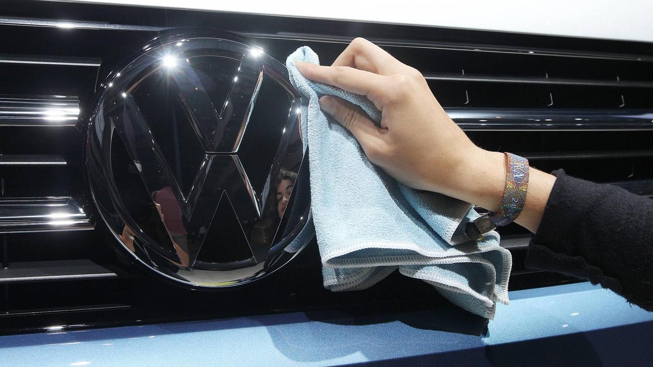 Eine Hand wischt mit einem Tuch über den Rand des VW-Logos an einem Auto.