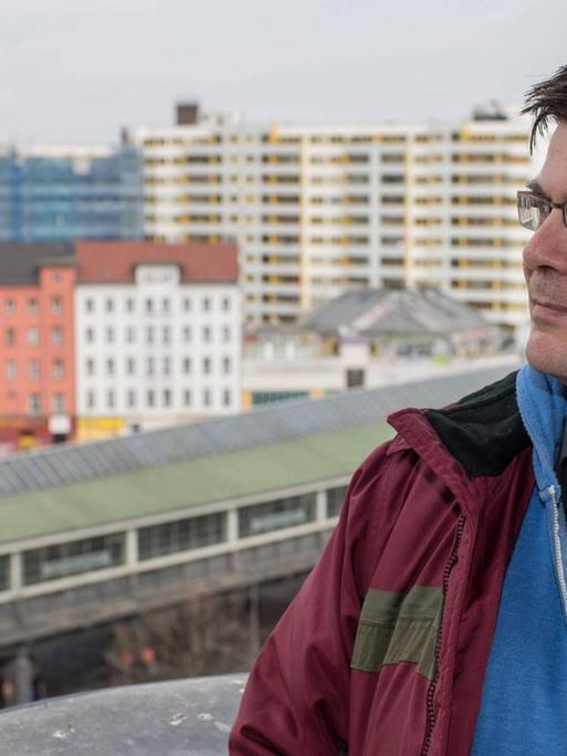 Andrej Holm steht auf einer Terrasse in Kreuzberg mit dem Wohnkomplex Zentrum Kreuzberg in seinem Rücken.