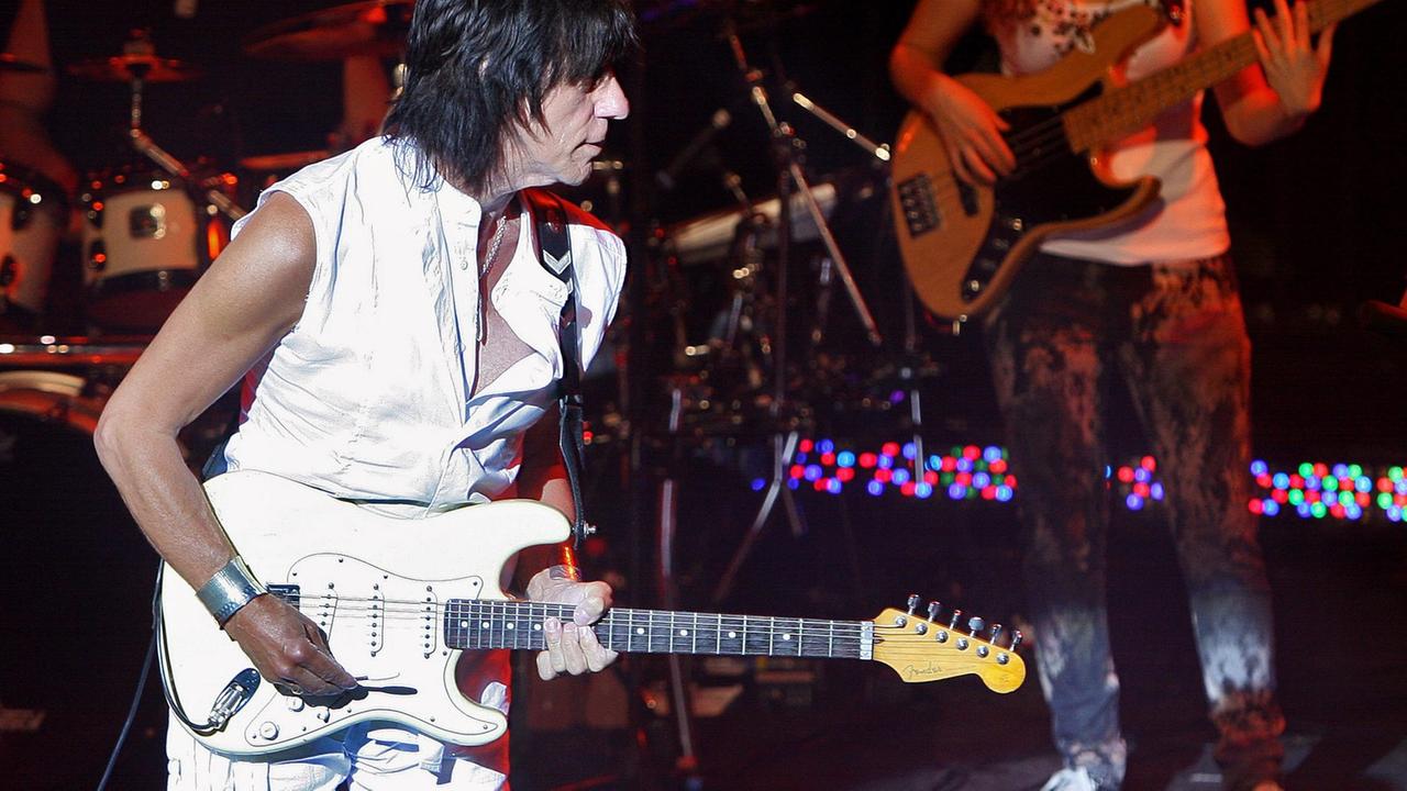 Ein in weiß gekleideter Mann spielt auf einer weißen Gitarre, im dunklen Bühnenhintergrund steht eins Frau mit Baßgitarre.