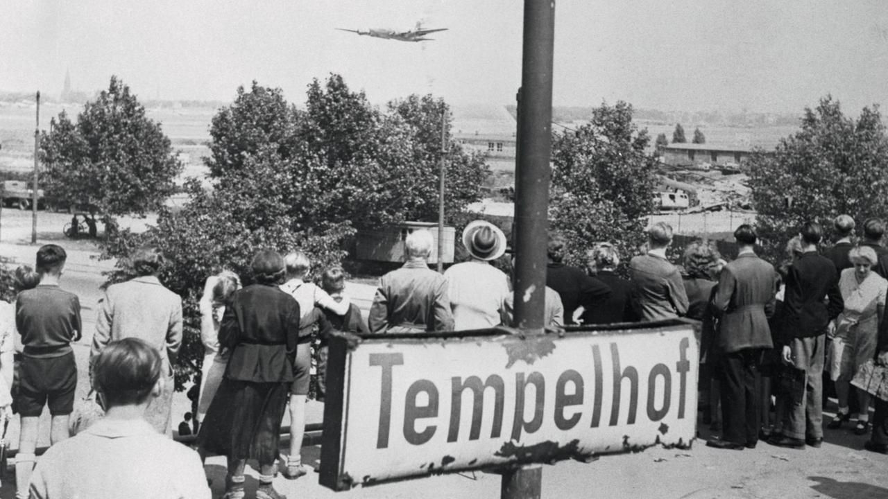 Passanten beobachten ein Flugzeug beim Landeanflug, im Vordergrund ein Schild mit der Aufschrift "Tempelhof".