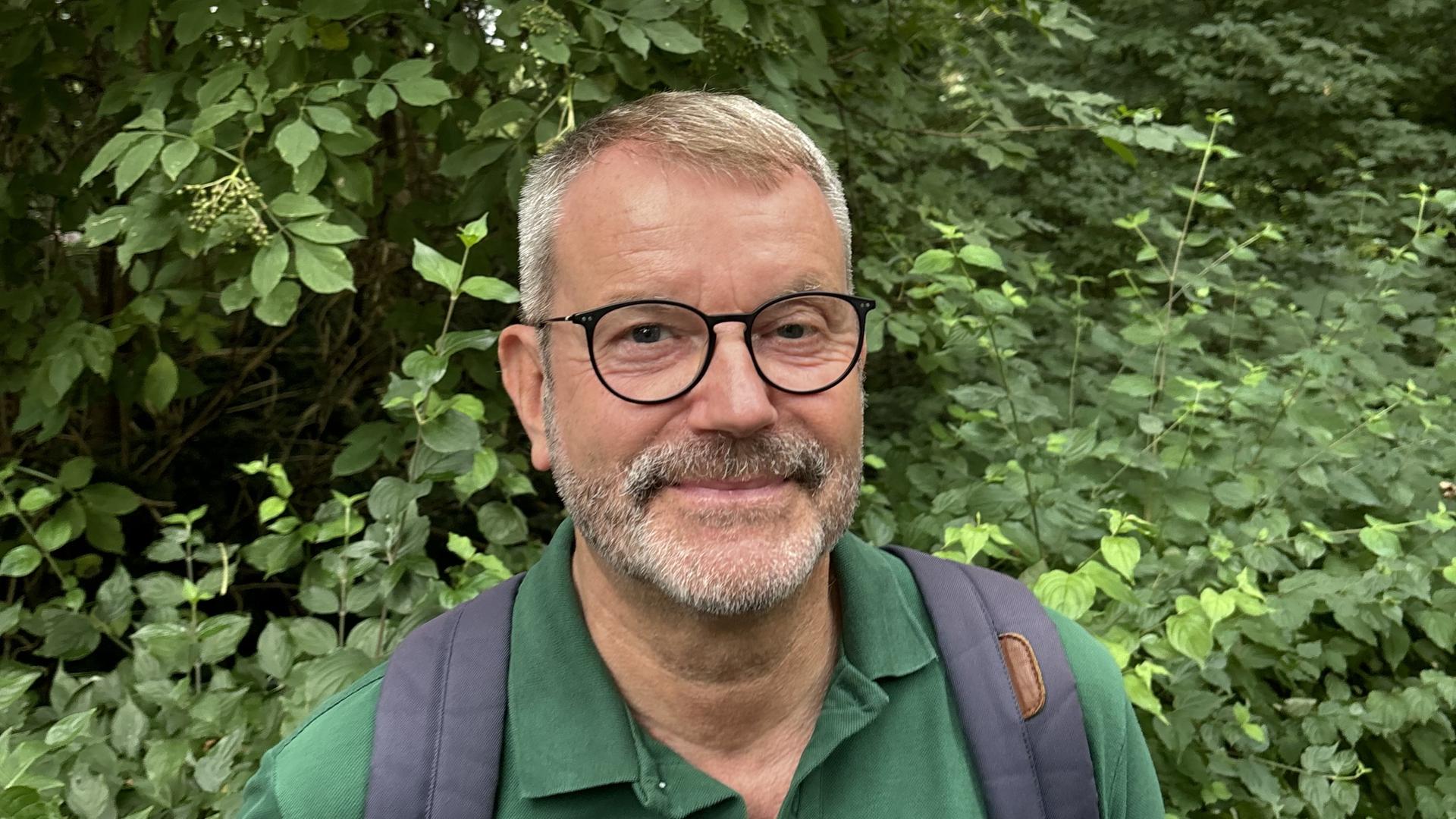 Bernd Brummer trägt eine schwarze Brille, ein grünes Poloshirt und einen Rucksack und steht vor einem begrünten Hintergrund in freier Natur