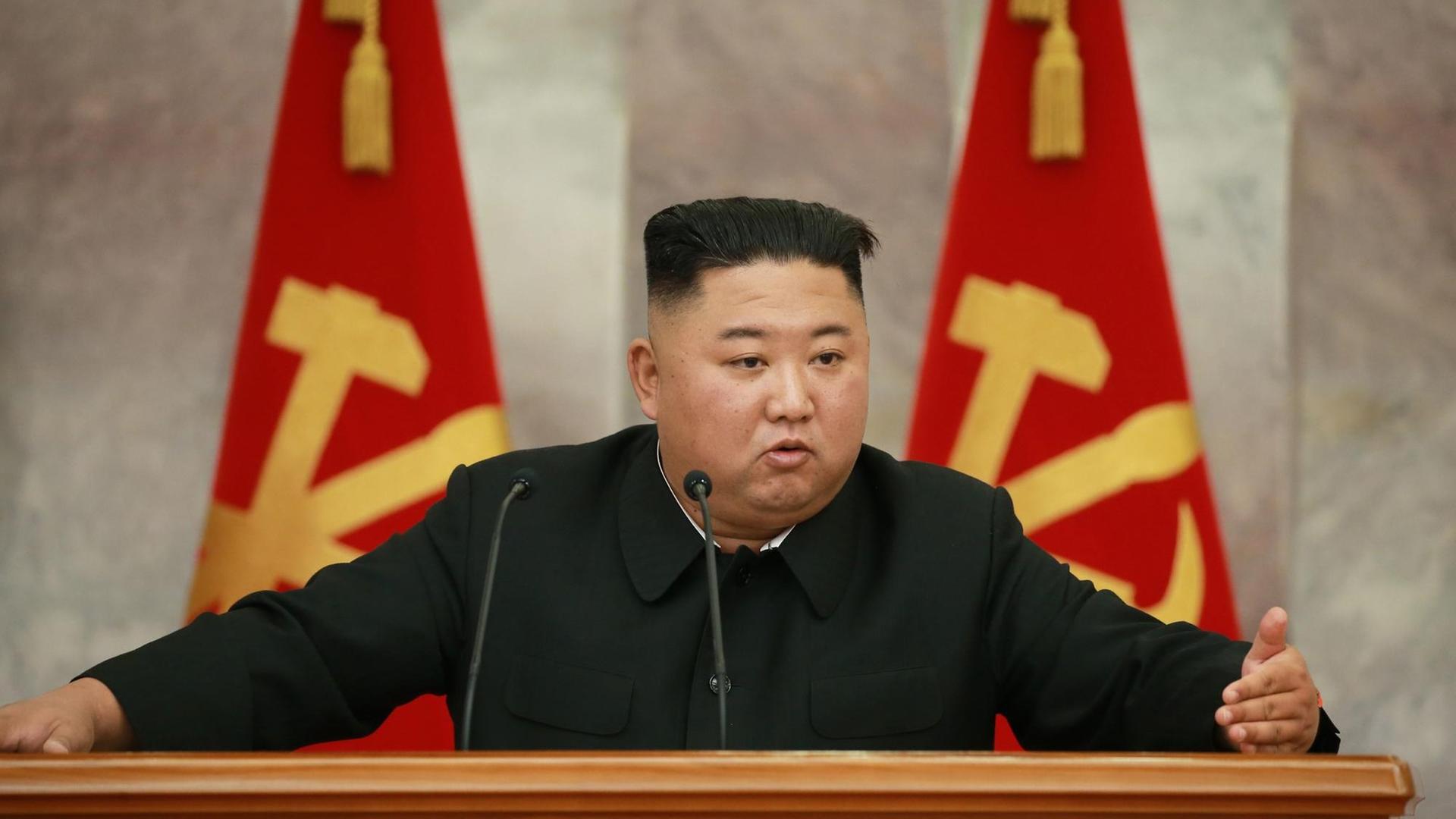 Kim Jong-un, gekleidet in Schwarz, steht an einem Rednerpult vor zwei Flaggen.